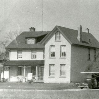 A black and white 1950s photograph of a two and a half storey house with a black car in the driveway. 