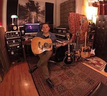 Ian Smith sitting with his legs crossed in a recording studio with a guitar in his hands