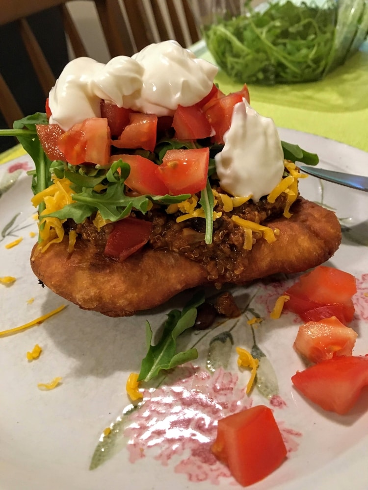 A piece of fry bread on a plate topped with chili, cheese, arugula, tomato and sour cream.