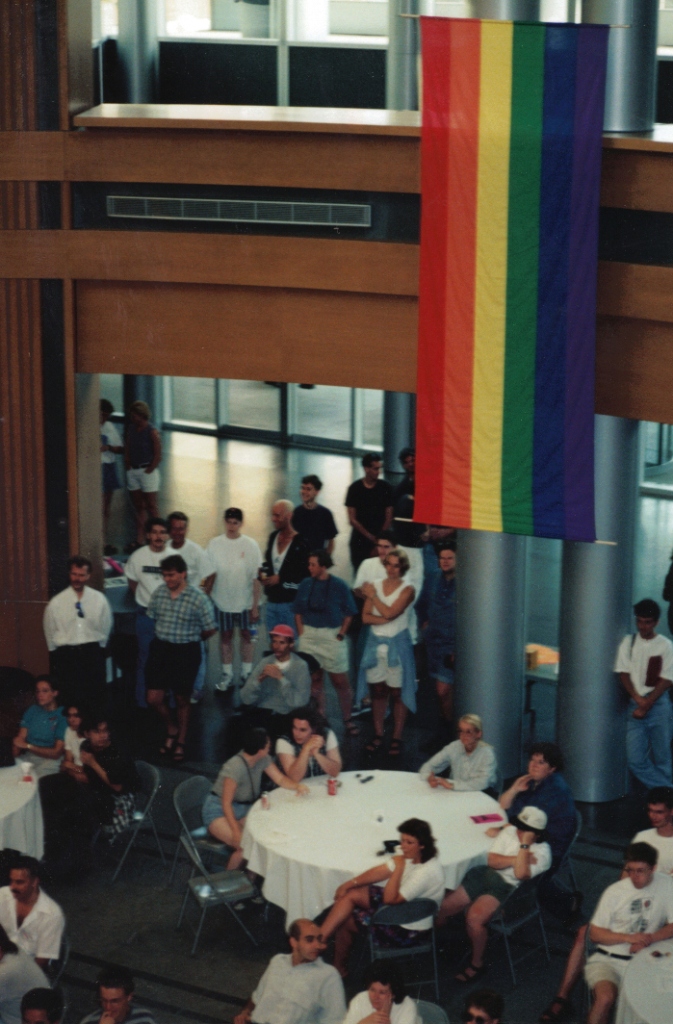 A photo taken from above, looking down at a crowd of people standing and sitting at tables at the Kitchener City Hall rotunda. A large rainbow flag hangs from the ceiling.