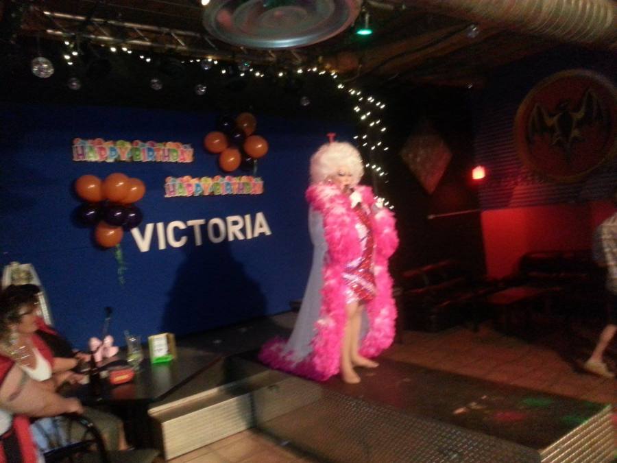 A photo of the inside of Club Renaissance. Drag queen Victoria Parks performs in a robe trimmed with a huge amount of pink feathers, a sparkly patterned dress and a fluffy white wig. Behind her at the back of the stage are banners that read Happy Birthday Victoria.