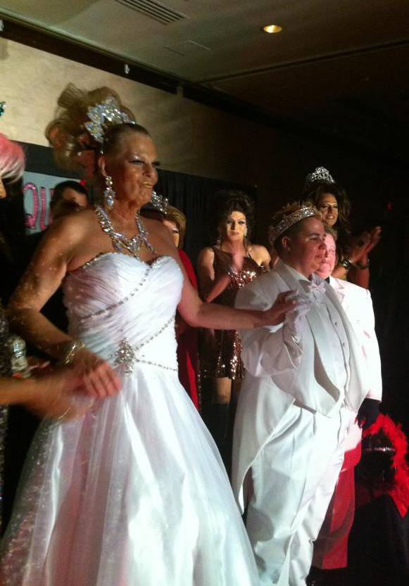 Late drag queen Robin Derring performs on stage in a gorgeous white gown, diamond jewelry, and a sky high blonde updo. Beside her is a drag kind in an all white tux and tails wearing a crown. Multiple drag performers stand behind them clapping.