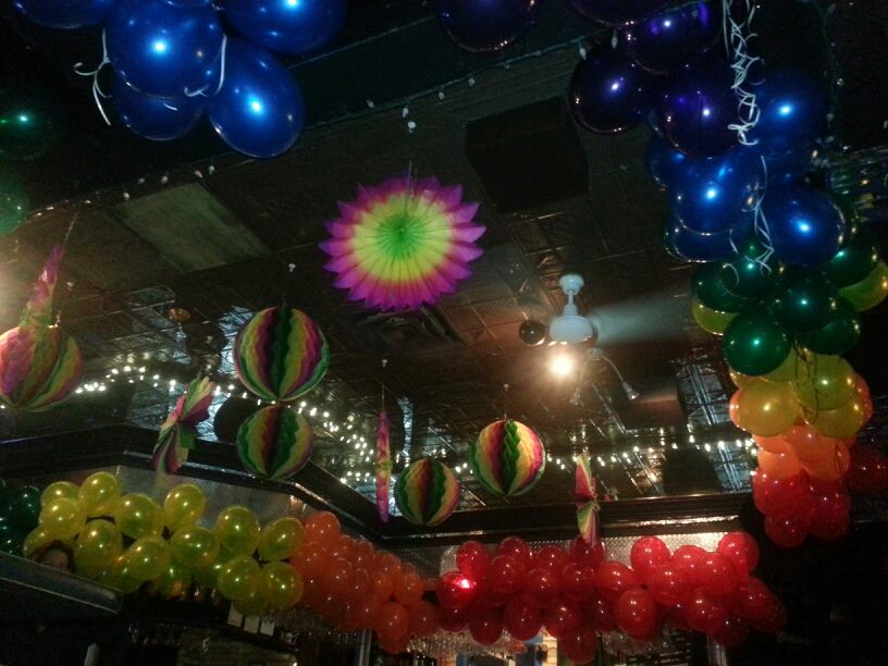 A photo of the ceiling around the bar at Club Renaissance. Surrounding the bar are balloons arranged by colour into a rainbow and from the ceiling hang multi-coloured paper decorations.