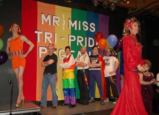 A photo from inside Club Renaissance. Miss Drew presides over the Mr and Miss TriPride pageant in a red gown and hairpiece. Seven drag kings and queens post on the stage in front of a large rainbow banner that reads "Mr and Miss TriPride Pageant".