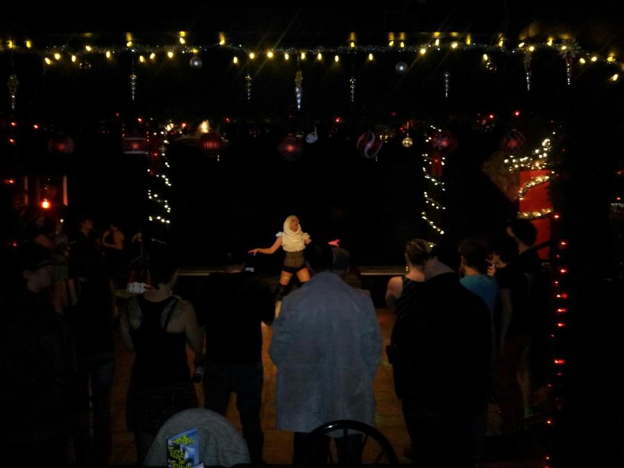 A photo from inside Club Renaissance. A crowd is gathered around the dance floor as a drag queen performs in the middle of the stage. Only string lights and one spotlight on the queen light the scene.