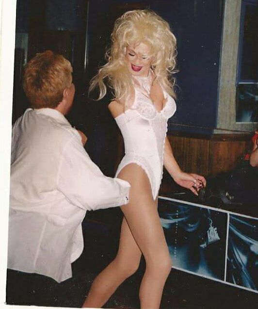 A photo of the gorgeous drag queen Miss Drew wearing a white lace bustier, red lipstick and a bouncy blonde wig. A woman with short blonde hair and a white dress shirt is dancing with her.