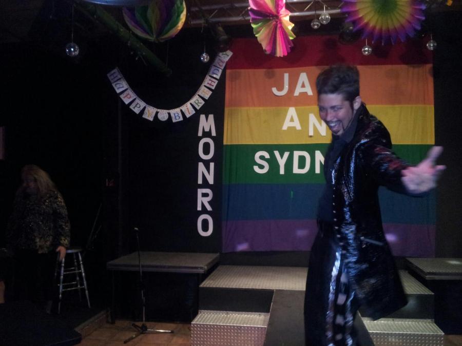 A photo of the stage and runway at Club Renaissance. A drag king in all black with a long coat and a goatee reaches out his hand to the audience. Behind him a pride flag hangs with the name Jay and Sydney. There are decorations hanging from the ceiling and beside the flag there is a banner that reads Happy Birthday and the name Monro.