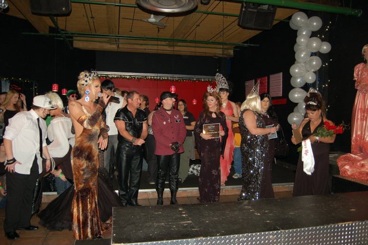 A photo of the inside of Club Renaissance during a drag pageant. Many drag queens and kings are standing beside the stage and runway wearing crowns. Miss Drew presides over the ceremonies with a microphone.