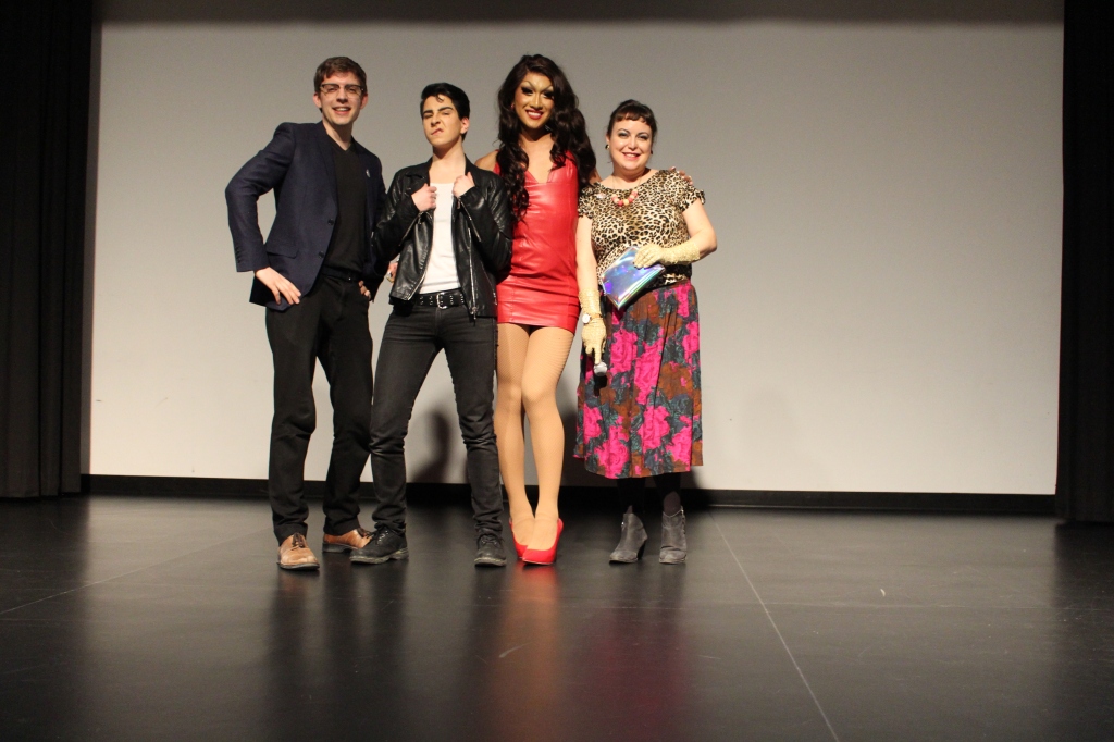 A photo from inside the Kitchener Public Library's theatre. Staff members Curtis Williams and Ellie Anglin pose on stage with drag king George Swooney and drag queen Kyne.