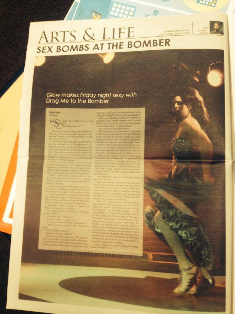 A photo of a newspaper open to a page with an article about the Drag Me To The Bomber shows held at the University of Waterloo. The headline reads "Glow makes Friday night sexy with Drag Me To The Bomber" and there is a large photo of a drag queen twirling under a spotlight.