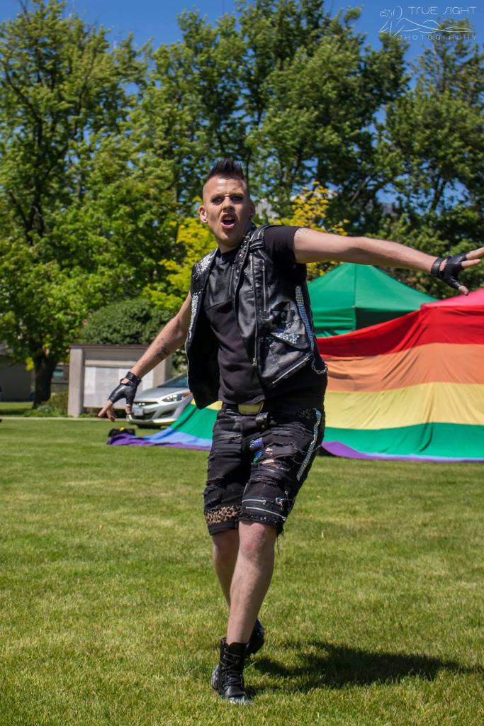 A photo of drag performer Troy Boy Parks performing in Victoria park during TriPride. Troy wears a leather vest and gloves and lip syncs dramatically in a grassy field in front of large pride flags.