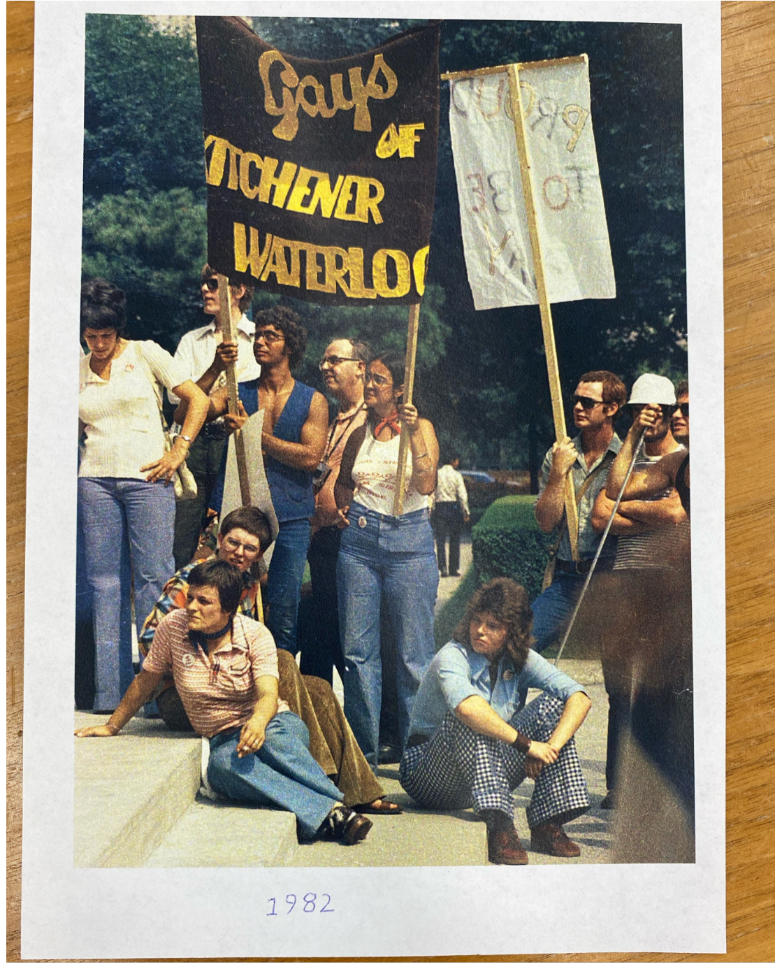 A photo of LGBTQ+ people demonstrating, two of them hold a yellow and brown banner that reads "Gays of Kitchener Waterloo", from 1982.