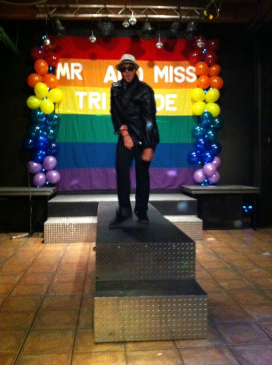 A photo from inside Club Renaissance. A drag king with a jacket, sunglasses and a fedora dances on stage. Behind him is a rainbow banner, surrounded by rainbow balloons, that reads "Mr and Miss TriPride"