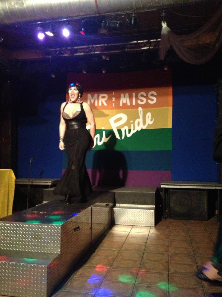 A photo from inside Club Renaissance of a drag queen in a black gown lip syncing powerfully to a song. Behind her at the back of the stage is a large rainbow banner that reads Mr and Miss TriPride.
