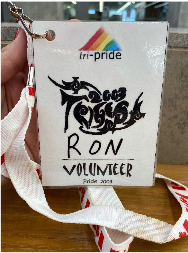 A photo of a laminated badge on a lanyard that reads" tri-pride" "Ron" "Volunteer" and "Pride 2003". The lanyard has Canadian flags on it.