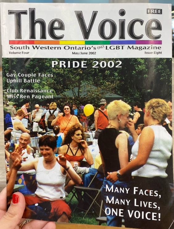 A photo of the cover of The Voice magazine from 2002. It featured a photo of Pride celebrations in Victoria park, including a crowd of people, a yellow balloon, Miss Drew and trees in the background.
