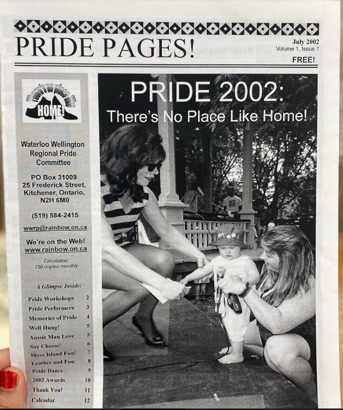 A photo of the cover of Pride Pages magazine, celebrating Pride 2002. The theme for that year was "There's No Place Like Home!" and the cover show a photo of a parent and baby interacting with a drag performer on the gazebo in Victoria Park.